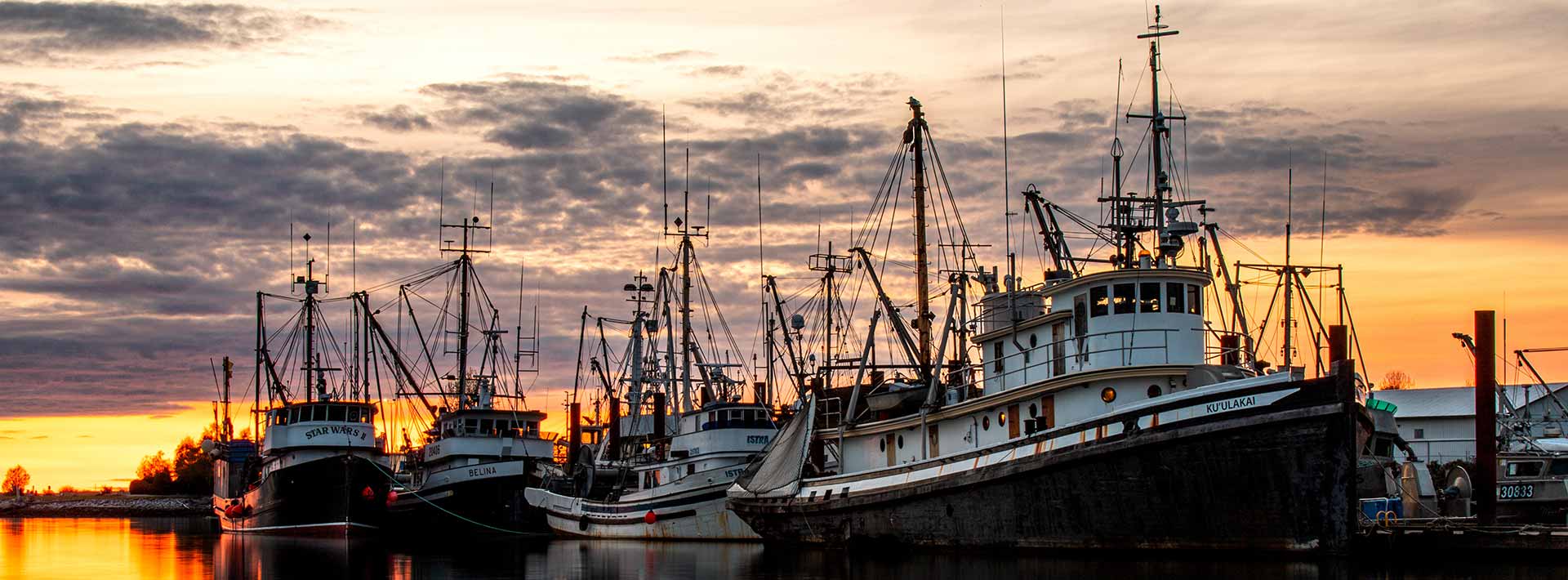 Boats rest peacefully along the banks of the Fraser River in Richmond, silhouetted against a breathtaking sunset backdrop