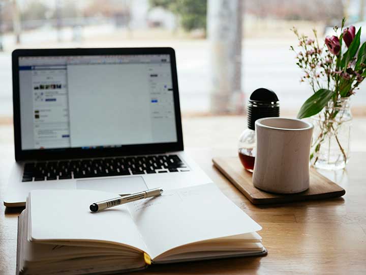An open notebook with a pen balanced across it, a blurred laptop in the background, both tools for generating ideas and asking questions.