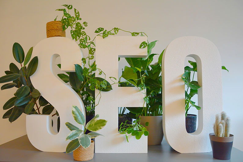 White SEO Sign on a Desk with Green Plants Wrapping Around it Kav’s Note: Sorry we can update the name of this to plants before uploading