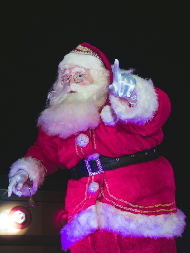 Santa Claus pointing his left index finger while holding a bell in his other hand.