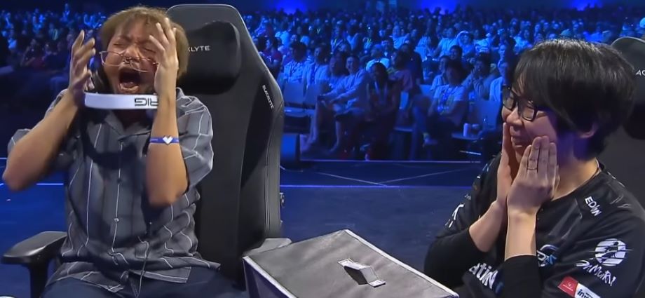 KojiKOG and Go1, two SNK fighting game players, on stage at EVO. Koji is pretending to have a meltdown post match, Go1 has his hands on his face.