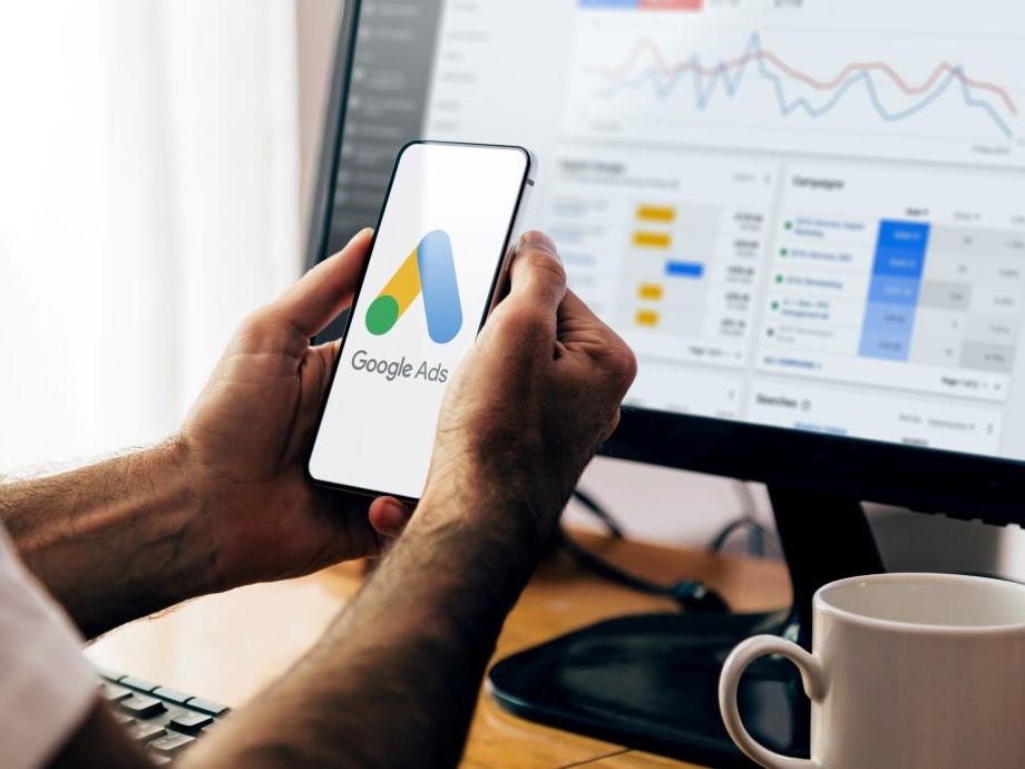 A man holding a mobile phone with the Google Ads icon on the screen and a desktop computer in the background with the Google Ads interface in soft focus.