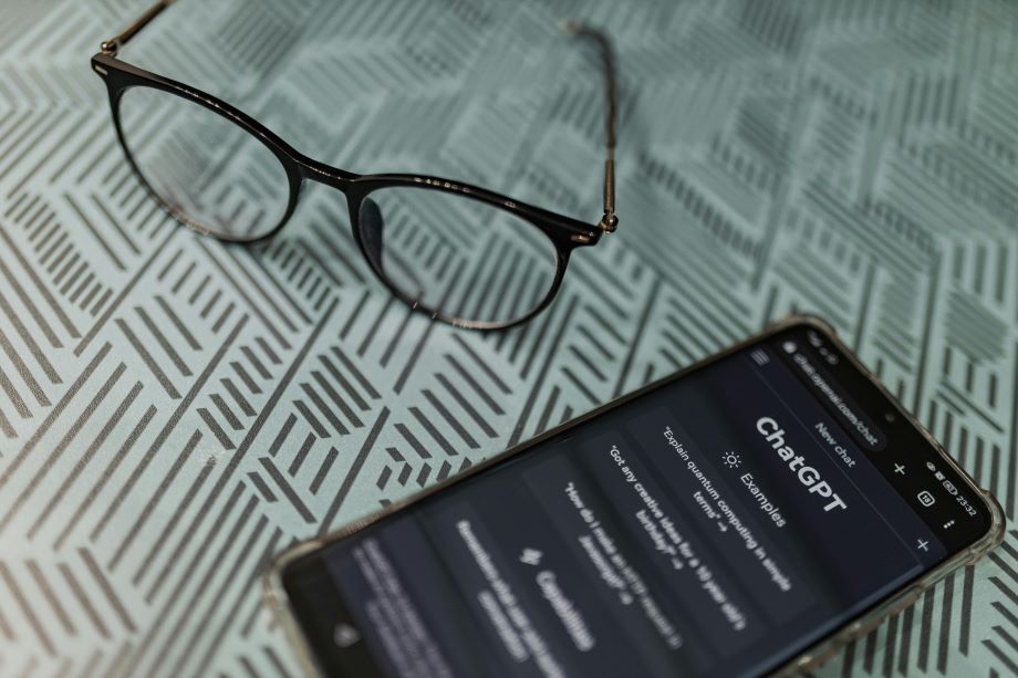 A smartphone showing the ChatGPT interface next to a pair of reading glasses on a patterned surface.