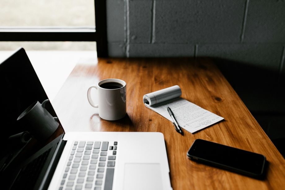 a tabletop with a laptop, phone, pen and notepad, and mug of coffee on top.