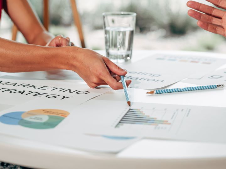 Decisions Close-up of a person pointing a blue striped pencil at printed marketing charts and documents on a white table.